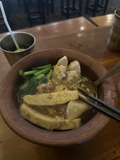 An overhead, tilted shot captures a meal served in a rustic, unglazed terracotta bowl, likely within an eatery in Kecamatan Cisaat, Indonesia. The bowl is filled with a dark broth containing yellow noodles, several large, light-colored meatballs, flat, elongated pieces (possibly fish cake or tofu) garnished with sesame seeds, green leafy vegetables (resembling bok choy or similar), and a lighter, possibly steamed dumpling or fish paste item. A metal spoon and black chopsticks are placed within the bowl, suggesting the meal is either about to be eaten or is in progress.
To the lower left, a metal cup with a white straw indicates a beverage. In the upper left background, part of another ceramic cup is visible, alongside what appears to be a wooden table surface and a dark, textured wall or partition. The overall lighting is dim, suggesting an indoor setting during the evening or with artificial light. No people are visible in the image, and no discernible text is present. The focus is entirely on the food and its immediate surroundings.