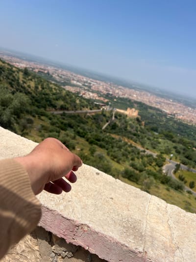 View from an elevated position overlooking Beni Mellal, Morocco. The image shows an urban landscape transitioning into dense greenery, with a clear blue sky above. A person's hand and part of their light brown sleeve are visible in the foreground, placed near a weathered, light-colored stone edge. The edge could be a wall or the side of a building. In the distance is a winding road amid the trees. The image suggests the photographer is on a high vantage point, potentially a building or hilltop, looking out over the city and surrounding landscape.