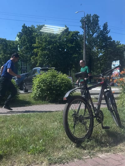 The image depicts an outdoor scene, likely in Barysaw, Belarus, on a sunny day. The primary focus is on a man in a green t-shirt standing next to a black bicycle. The bicycle is a prominent object, with its frame, wheels, and handlebars clearly visible. The man appears to be interacting with the bicycle, possibly in the process of working on it or preparing to ride.

In the background, lush green trees suggest a temperate season. A light pole with a streetlamp is also visible, indicating an urban or suburban setting. Further in the distance, a dark-colored car is parked near a building with green window frames. Another person, wearing a blue t-shirt and black pants, is seen in the lower part of the image, facing away from the camera and appearing to be walking.

A paved pathway runs alongside the grassy area where the bicycle and the man in green are located. There is a sign in the background with some text, but it is not clearly legible due to its distance and resolution. The overall impression is of a casual daytime activity taking place outdoors. The bright sunlight and clear blue sky indicate good weather conditions. The presence of trees, a streetlamp, and a car suggests a location within a town or city.