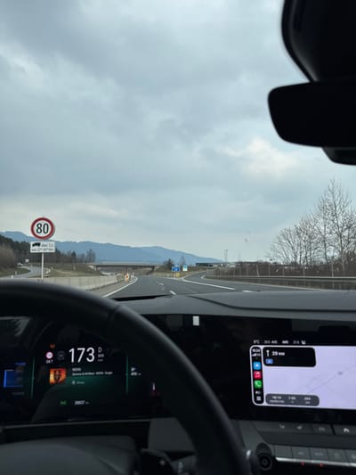 The scene is viewed from inside a vehicle on a multi-lane highway near Aichdorf, Austria, under an overcast sky. The lower left of the frame is occupied by a dark steering wheel. The digital instrument cluster behind it displays a speed of 173 km/h, 'D' for drive mode, 'ECO' indicator, and an odometer reading of 38,837 km. An album cover for "MOVA" by "dannn & M.M.Run" is visible, indicating music playback from an iPhone. To the right, an infotainment screen shows a navigation map with 29 km remaining to the destination, the current time as 16:24, and an outside temperature of 8°C. On the left side of the highway, a circular speed limit sign displaying "80" is present, with a supplementary sign stating "über 7.5t von 22-05 Uhr" (for vehicles over 7.5 tons from 10 PM to 5 AM). The highway, marked with white lines, extends into the distance, passing beneath a concrete overpass. Distant mountains are visible on the horizon, and bare deciduous trees line the right side of the road. No individuals are visible.