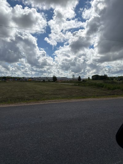 The image depicts an outdoor scene in Jaunsaurieši, Latvia, under a partly cloudy sky. In the foreground, a dark asphalt road occupies the bottom half of the frame, with a sliver of its shoulder visible along the top edge. Beyond the road lies a large, mown grassy field. In the mid-ground, a long, low-rise building with a white facade stretches horizontally across the landscape. In front of the building, numerous parked trucks and trailers are visible, suggesting a commercial or industrial area. A line of trees forms a dark green border behind the building, and further back, more trees and vegetation are present. The sky is filled with dramatic, textured clouds, with patches of blue sky visible between them. There are no people or animals visible in the image. The overall impression is one of an industrial facility situated in a rural or semi-rural setting.