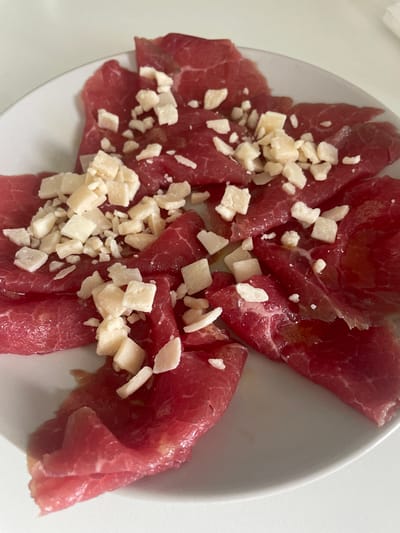 A white plate of thinly sliced carpaccio topped with pieces of Parmesan cheese sits on a white surface. The carpaccio is a deep red color, and the cheese is white. The location is not apparent from the image.