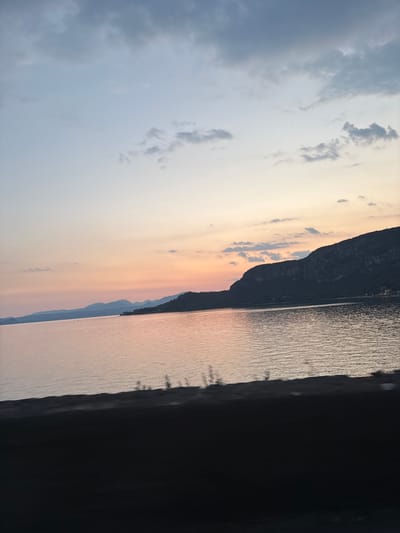 A scenic view captures a tranquil lake reflecting the soft hues of a sunset. In the distance, a silhouette of a mountain range borders the horizon, its peaks softened by the fading light. The sky above transitions from a pale blue at the top to a warm gradient of pink and orange near the horizon, dotted with scattered clouds. The foreground is dominated by a dark, out-of-focus expanse, likely a roadside barrier or embankment, obscuring the immediate surroundings. The scene evokes a sense of peace and natural beauty, characteristic of the Lake Garda region.
