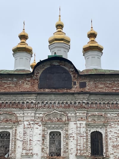 The image shows a close-up view of the upper facade and domes of the Church of the Ascension in the village of Kstinino, Tver Oblast, Russia. The church features multiple golden domes with crosses atop white octagonal drums. The brickwork of the building is weathered and in a state of disrepair, with peeling white plaster revealing the red brick beneath. Decorative brickwork and window surrounds are visible. The sky is overcast and grey.