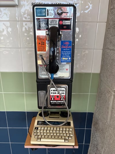 A payphone is mounted on a tiled wall in North East Township, United States of America. The payphone is black with chrome accents and features a handset, a coin slot, and various labels with pricing and instructions. Below the payphone, a beige keyboard with a black cord is placed on a wooden shelf. The wall is tiled with a pattern of white and light green tiles, and a section of dark blue tiles is visible at the bottom. A rough, grey concrete block wall is to the right of the payphone. The scene appears to be indoors, possibly in a public building or transit station.