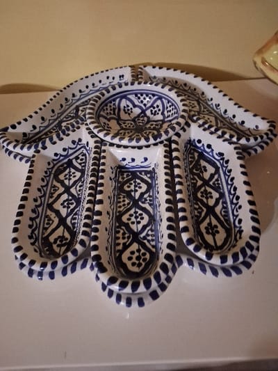 A white, ceramic serving dish with cobalt blue designs rests on a surface. The dish is shaped like a hand with five distinct compartments. The central compartment is round, while the other four are elongated ovals. Each section features intricate patterns and designs in the blue color, contrasting with the white ceramic base. The edges of the dish are decorated with small, evenly spaced dots, adding to the overall artistic detail.