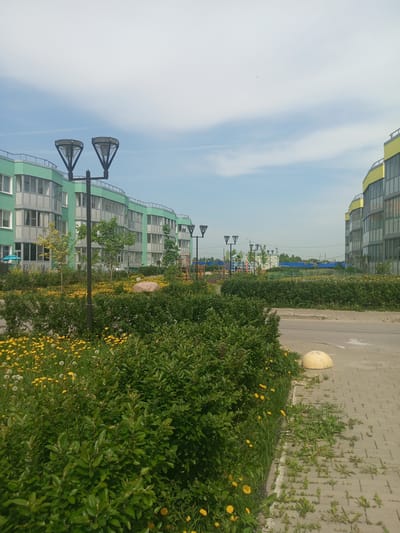 The image depicts a street scene in Kommunar, Russia. In the foreground, there is a paved sidewalk adjacent to a street. A hemispherical concrete barrier sits on the edge of the sidewalk. Lush green bushes and a field of dandelions are growing along the sidewalk.
In the background, modern apartment buildings can be seen. The buildings are predominantly green and yellow in color. There are ornamental street lamps visible in the middle ground. A playground with brightly colored equipment is situated between the buildings. The sky is light blue with scattered clouds.