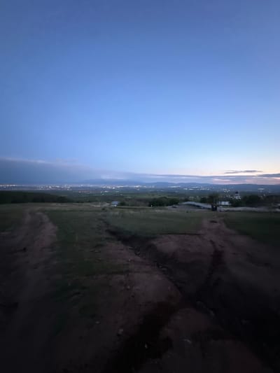 The image displays a twilight landscape scene, likely captured from an elevated viewpoint overlooking a valley or plain in Dobroslavtsi, Bulgaria. The sky occupies a significant portion of the upper left, transitioning from a deep blue at the top to lighter blues and faint hints of pink/orange along the distant horizon, characteristic of dusk or early evening. A few wispy clouds are visible near the horizon line.
Below the horizon, distant mountains or hills are silhouetted against the fading light. The expansive midground is a vast stretch of land, possibly a valley, dotted with numerous small, twinkling lights, indicating scattered settlements, villages, or a town. A dark, meandering line, which could be a road or a river, can be faintly seen winding through the illuminated area.
The immediate foreground, occupying the right side of the image (when oriented horizontally), is dark and indistinct, showing uneven ground with minimal detail, suggesting a natural, uncultivated terrain with sparse vegetation or dirt. There are no identifiable human activities or specific objects in this dark foreground.
No people or explicit events are visible. The overall mood is one of quiet vastness and peaceful transition as daylight fades and artificial lights begin to punctuate the landscape. There is no visible text in the image.
