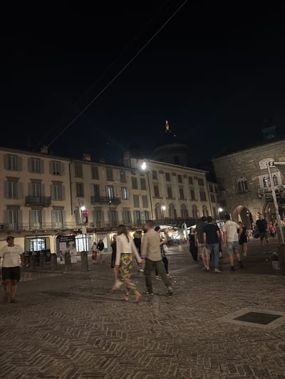 The image depicts a nighttime scene in Città Alta, Bergamo, Italy, specifically the Piazza Vecchia. The foreground features a cobblestone street with several individuals walking. To the left, a man in a white t-shirt and dark shorts is walking towards the viewer. In the center, a couple walks away from the viewer, the woman in a white top and patterned pants, the man in a light-colored shirt and dark pants. Behind them, a group of people are walking in various directions, including a man in a white t-shirt and grey shorts. The background is dominated by historic buildings. On the left is a building with cream-colored facade, multiple windows with closed grey shutters, and balconies. Below the windows, a sign reads "CREDITO BERGAMASCO". To the right of this building is a dome topped with a golden statue, a prominent feature of Città Alta. Further to the right is a stone building with arched windows and a clock tower. Streetlights illuminate the scene, casting shadows on the cobblestones and highlighting the architecture. The atmosphere is that of a lively evening in a historic Italian city.