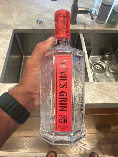 An indoor scene depicts a person's right hand holding a clear glass bottle of "DEVIL'S GRIN TEXAS GIN" vertically in the foreground. The bottle has a red label with white text, stating "750 ML | 44% ALC/VOL | 88 PROOF", and features embossed floral or vine patterns on its sides. The bottle's cap is red foil with black illustrations of a globe and a "DG" logo. The person's left foot, wearing a dark sandal, is visible at the bottom of the frame, and a dark, possibly olive green, fabric wristband or watch strap is visible on their right wrist.
The background shows a light-colored, mottled granite or quartz countertop with a double-basin stainless steel kitchen sink. A silver-colored kitchen faucet is positioned between the two basins. The right sink basin contains a metal strainer and what appears to be a clear glass or plastic cup. To the right of the faucet on the countertop are two pump bottles of cleaning or soap product: a purple bottle labeled "MRS. MEYER'S" and a green bottle specifically labeled "MRS. MEYER'S CLEAN DAY HAND SOAP." The floor is visible in the lower portion of the image, consisting of light-colored, horizontal wood planks. The environment is consistent with a domestic kitchen setting within the United States, but no specific visual markers identify the location as Aspen.
