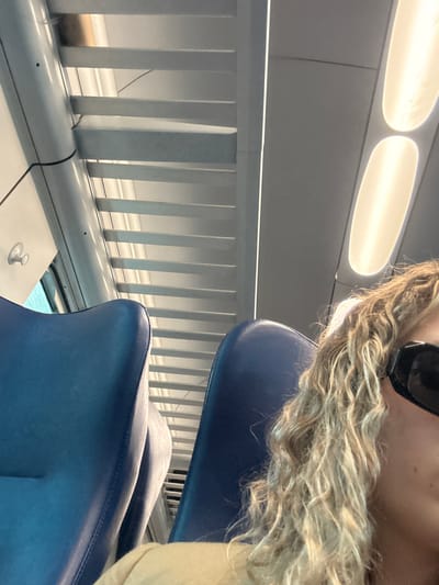 The image shows a person with blonde, curly hair wearing dark glasses, sitting in a blue seat on what appears to be a train or bus. A white railing with vertical bars is visible in the background, next to a white wall. There are two bright, elongated lights on the ceiling. There is no verifiable information present in the image to confirm the specific location, Alessandria, Italy.