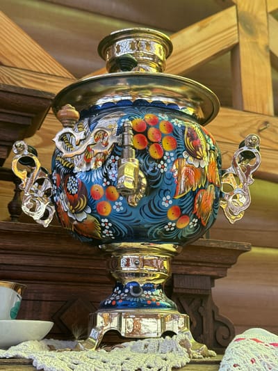 The image shows a Russian samovar, elaborately decorated with a blue background, red berries, and orange bird motifs. It features ornate silver handles and spigot. The samovar is positioned indoors, likely in a room with wooden walls and possibly part of a display or exhibition in the city of Kladovitsy, Russia. A white, crocheted object is visible on the right side of the image.