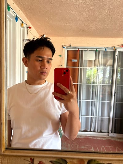 An adult male with dark, spiked hair, a faded haircut, and a silver stud earring in his left ear is depicted taking a mirror selfie. He wears a white crew-neck t-shirt and a thin gold chain. A gold-toned ring is present on the fourth finger of his left hand, which holds a red smartphone featuring a dual-camera system. The background, reflected in the mirror, shows a textured, light peach-colored ceiling or soffit. Below it, a string of multi-colored string lights (red, green, blue, yellow) is visible. Behind the subject are white-framed sliding glass doors or windows with a grid pattern, reflecting outdoor greenery, including what appears to be palm trees. The bottom edge of the mirror reflection shows a reddish-brown surface with faded white text. The specific location within Thousand Oaks, United States, cannot be determined from the visual information provided.