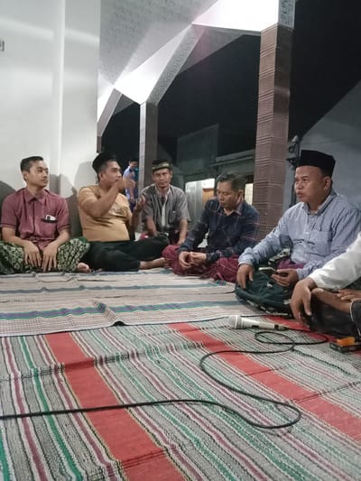 In Brangsong, Indonesia, a group of men are gathered indoors, seated on colorful woven mats. The room appears to be a communal space, possibly a mosque or a community hall, with a distinctive textured ceiling and large structural pillars. Five men are clearly visible in the foreground, all dressed in traditional Indonesian attire, including sarongs. One man in a red collared shirt sits on the far left, facing forward. Next to him, a man in a tan t-shirt is gesturing with his hands while speaking. To his right, a man in a gray shirt is looking down. Further to the right, a man in a blue checkered shirt is holding a smartphone and looking to his left. The man closest to the camera on the right wears a black peci (cap) and a blue and white checkered shirt. A microphone and its cable are laid out on the mat in front of the seated men, suggesting a presentation or discussion is taking place. Another man is partially visible on the far right, with his hand resting on his lap. In the background, a few other figures are present, including a young boy in a blue shirt. The lighting suggests it is evening or nighttime, with artificial light illuminating the gathering.