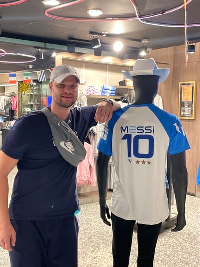In a retail environment, possibly within Aeropuerto Internacional Ezeiza, Argentina, a man wearing a navy blue t-shirt, navy blue pants, a gray baseball cap, and a gray Nike fanny pack positioned across his chest, stands to the left of a black mannequin. The mannequin is dressed in a white and blue jersey with "MESSI" and the number "10" prominently displayed on the back, along with three gold stars. The mannequin also wears a light blue and white cowboy hat. The background shows shelves stocked with clothing and merchandise, and flags from various nations are visible on the wall, including Argentina, France, and Uruguay. The lighting suggests an indoor retail space.
