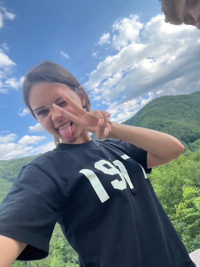 A young woman with dark hair, wearing a black t-shirt with the number "1997" printed in white, is taking a selfie outdoors. She is sticking her tongue out and making a peace sign with her right hand. The background consists of a lush green mountain range under a blue sky with scattered clouds. The location is identified as Pechinska, Bulgaria.