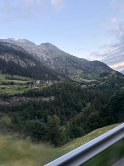 The image captures a panoramic view of a mountainous valley in Sainte-Foy-Tarentaise, France. The landscape is dominated by rugged, imposing mountains with a mix of rocky outcrops and dense coniferous forests. In the mid-ground, a quaint village with scattered buildings is nestled within the valley, surrounded by verdant green slopes and fields. A thin waterfall cascades down the side of a mountain in the distance. The sky above is a soft blue, with a few wispy clouds hinting at the approaching dusk or dawn. In the foreground, a metal railing, likely part of a viewing platform or roadside barrier, is visible, suggesting the photo was taken from an elevated perspective, possibly from a moving vehicle or a scenic overlook. The overall impression is one of serene natural beauty and rural charm.