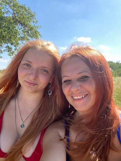 This is a close-up, outdoor selfie featuring two young women with red hair, positioned closely together and smiling at the camera. The woman in the foreground has fair skin and blue eyes, and is wearing a red bikini top and silver necklaces. The woman behind her has a wider smile and appears to be wearing a blue bikini top. Both women have long, wavy red hair that frames their faces.
The scene is bathed in bright sunlight, suggesting it's a sunny day, likely midday or afternoon given the high angle of the sun and clear sky. The background consists of a bright blue sky with some white clouds, and out-of-focus greenery which could be trees or tall grass, indicative of an outdoor, natural setting. There are no visible objects other than the jewelry worn by the women.
The overall mood of the image is joyful and relaxed, with the women appearing happy and enjoying themselves. The orientation of the photo is such that the subjects are tilted, as if captured in a spontaneous moment. The location context of Kyr'yakivka, Ukraine, suggests a possible holiday or leisure activity in a rural or coastal area during warmer weather. There is no visible text in the image.