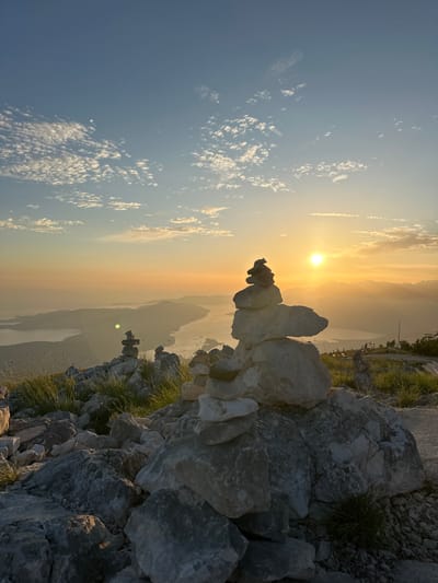 An outdoor, horizontal, medium shot captures a sunset over a bay, as viewed from a rocky mountain summit. Two cairns, or stacked stone markers, are in the foreground, with the larger one to the right of center. The cairn on the right is constructed of at least seven light-colored stones, varying in size and shape, stacked precariously. The cairn to the left is smaller and further in the distance. The rocky terrain is covered with patches of green grass and some dry, tall grass. The sun, a bright, yellow orb, is low in the sky, casting a warm, orange glow over the scene and illuminating a hazy mountain range in the distance. The sky is a gradient of blue and orange, with scattered white clouds. The bay below is vast and calm, with islands dotting its surface. The overall mood of the image is serene and majestic. The image appears to have been taken from a vantage point on a mountain overlooking the Bay of Kotor, in or near Mirac, Montenegro, a region known for its dramatic coastal scenery and hiking trails. There are no people or animals visible in the image.
