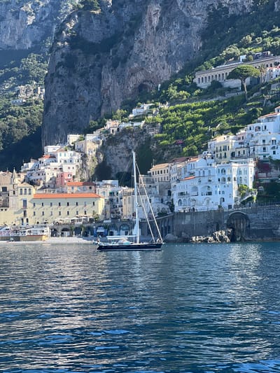 The image depicts the coastal town of Pogerola, Italy, as viewed from the water. Dominating the scene are steep, verdant cliffs that rise sharply behind a cluster of white and pastel-colored buildings. These structures cascade down the hillside, interspersed with lush greenery and terraced gardens. A prominent sailboat with white sails is moored in the foreground, its dark hull resting on the blue, rippling water. The coastline features a narrow beach with scattered umbrellas and a few sunbathers, suggesting a calm, sunny day. Stone walls and arched structures are visible along the shoreline, indicating historical architecture. The overall atmosphere is serene and picturesque, capturing the quintessential charm of the Amalfi Coast.