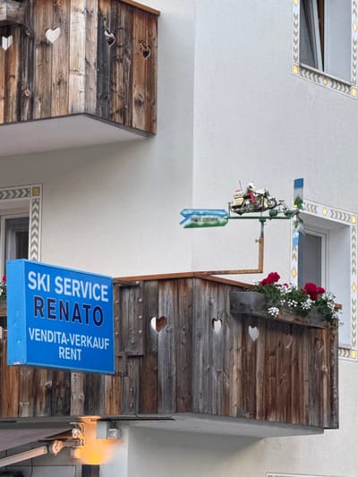 The image shows a close-up view of a balcony on a building in La Ila - Stern - La Villa, Italy. The balcony features a rustic wooden railing with heart-shaped cutouts. A blue sign is prominently displayed, reading "SKI SERVICE RENATO VENDITA-VERKAUF RENT". Above the balcony, a wind vane shaped like a motorcycle rider is visible. Flower boxes with red and white flowers adorn the balcony railing. The building exterior is painted white, and decorative geometric patterns are present around a window. A light fixture is visible on the underside of the balcony.