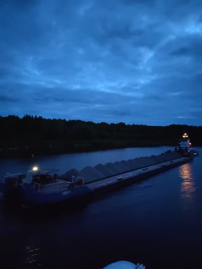 A barge is navigating a dark river or canal, likely the Volga River or a connected waterway, near Alexandrovskoye, Russia. The barge is heavily laden, possibly with sand or gravel, and lit by navigation lights. The surrounding area appears to be a forested landscape under a cloudy, twilight sky. The time of day is likely late evening or night.