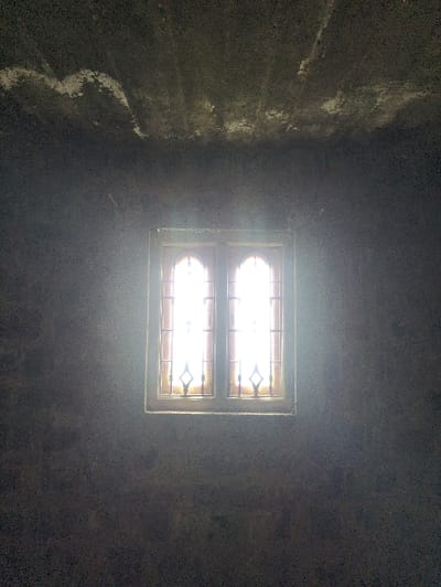 The image depicts an interior view of a dimly lit room in Kadaladi, India, focused on a brightly illuminated window. The window, positioned centrally, consists of two vertical panes set within a dark frame. Each pane features decorative metal security bars arranged in a diamond pattern with curved elements. Intense white light from outside the window obscures the exterior view, and visible light rays penetrate the dusty interior atmosphere. The surrounding walls and ceiling are dark grey to black, exhibiting a rough, textured surface. Irregular white patches, consistent with discoloration or material damage, are visible on the upper wall and ceiling. No individuals or specific actions are discernible within the frame beyond the interaction of light and dust.