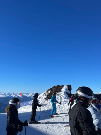 The image captures a vibrant, sunny day on a snow-covered mountain ski slope in Séez, France, showing at least five skiers paused amidst a scenic alpine landscape.
In the foreground, a skier in a black jacket and a black helmet with white goggles, featuring "SAVOIE" text on the strap, is visible from behind, holding ski poles. Another skier in a dark jacket and helmet is positioned just behind them. Further into the mid-ground, a skier in a striking bright blue ski suit and a yellow helmet stands on skis, situated next to a rugged, snow-covered rocky outcrop. Additional skiers are partially visible in the background.
The setting is a high-altitude ski resort, characterized by well-groomed snow, ski equipment, and resort infrastructure. A red marker post with the number "10" is visible to the left of the central group of skiers, and a blue banner with partially legible text (possibly "PST E") can be seen behind them. The background features expansive views of distant, snow-capped mountain ranges under a perfectly clear, deep blue sky, indicating excellent weather and visibility. The bright sunshine suggests it is likely midday or early afternoon. The "SAVOIE" branding on the goggles serves as a clear geographical cue, confirming the location within the Savoie region of France. The scene depicts a serene and active day of winter sports.