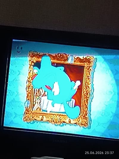 The image displays a screen, likely a television or monitor, showing a digital drawing. In the upper left corner of the screen, there is a small, simple smiley face with two dots for eyes and a curved line for a mouth. Dominating the center of the screen is a framed picture. The frame is ornate and golden, with detailed carvings. Inside the frame, a large, turquoise-colored cartoon character is depicted. This character appears to be a stylized fish or sea creature, with a large head, prominent fins, and an open mouth with a visible tongue. The character is posed in a dynamic manner, appearing to be swimming or leaping. The background within the frame is a reddish-brown color. The overall scene on the screen has a bright, blueish tint, suggesting lighting effects or a specific color filter applied. Below the screen, a timestamp is visible in the lower right corner, reading "25.06.2026 23:37". There is no information or visual cue within the image that relates to the city of Bolhrad, Ukraine.