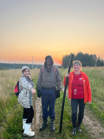 Three individuals are standing on a dirt path in a rural field during sunset. The person on the left, a woman, is wearing a camouflage hooded jacket over a grey top, black leggings, and white rubber boots. She has a red and black backpack and is holding a wooden stick. The person in the center, a man, is wearing a grey hooded jacket with a mesh covering over his face, blue pants, and black boots. He has a grey backpack. The person on the right, a woman, is wearing an orange hooded jacket over a red t-shirt, patterned leggings, and grey and black shoes. She has a yellow and green bandana on her head and is holding a long black pole. The background consists of a grassy field with trees in the distance, and the sky is a gradient of pale blue and orange, indicating sunset. The location is identified as Badashka, Russia.