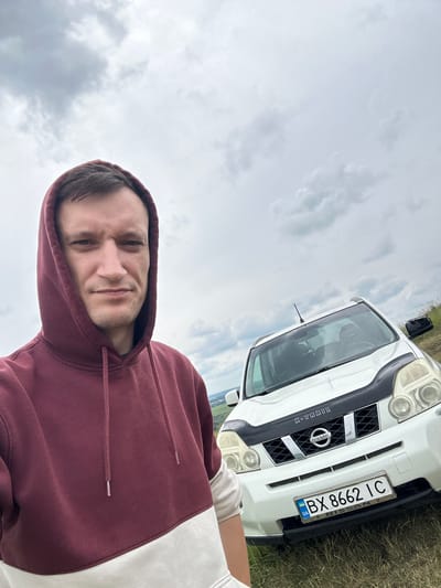 A man wearing a maroon and white hoodie stands in front of a white Nissan X-Trail SUV. He is facing the camera and appears to be taking a selfie. The car has a license plate that reads "BX 8662 IC". The background is an overcast sky with clouds. The scene is outdoors, on a grassy field, possibly in the city of Humentsi, Ukraine, based on the license plate.