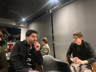 Several young men sit in a dim room with dark grey walls. One man in the foreground wears a black jacket and holds a cigarette to his lips. Another man sits to the right in a black jacket and patterned pants, gazing off to the side. In the background, a TV screen reflects another man and there is a bottle of Coca-Cola visible on the table. Artificial light fixtures shine from the ceiling.