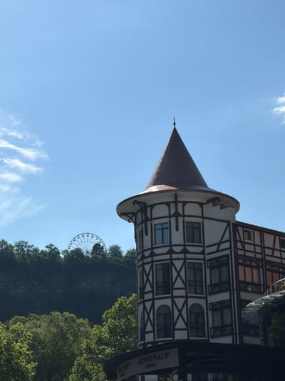 The image shows a view of the Crowne Plaza Borjomi hotel in Borjomi, Georgia. In the foreground, the distinctive architecture of the hotel's tower is visible, featuring a conical roof and half-timbered detailing. The hotel is situated amidst lush green trees. In the background, a ferris wheel is visible on top of a tree-covered hill, suggesting a recreational area or amusement park. The sky is clear and blue with a few wispy clouds. The scene appears to be a bright, sunny day.