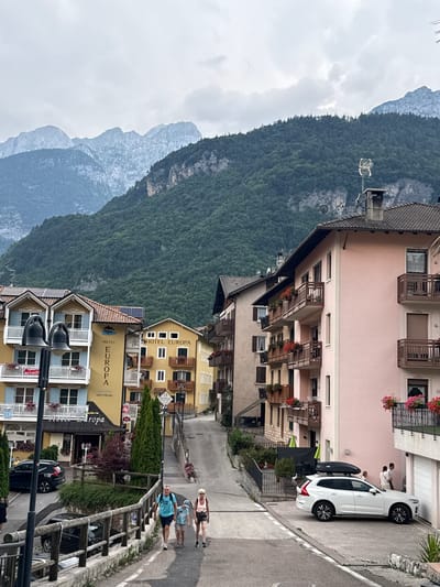 The image depicts a street scene in Molveno, Italy, dominated by a mountainous backdrop and buildings lining the street. The foreground features a paved road with a few people walking and a white SUV parked on the right side. A wooden railing runs along the side of the road. Several buildings are visible, including hotels and residences, with balconies adorned with plants. The buildings are of varying colors, with a prominent pink building on the right and a yellow building labeled "HOTEL EUROPA" on the left. The background is characterized by lush green mountains under a cloudy sky. A few streetlights are also present in the image.