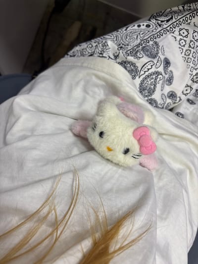 A fluffy white Hello Kitty plush toy with pink accents is lying on a crumpled white sheet. The toy is positioned diagonally, with its head facing towards the left side of the frame. Its eyes are black dots, and its nose is a small orange triangle. A pink bow is attached to the right side of its head. In the foreground, several strands of blonde hair are visible on the sheet. Behind the toy and the sheet, a patterned fabric with black and white paisley designs is partially visible. The lighting suggests an indoor environment. The scene does not contain any discernible elements that would place it specifically in the city of Kuznetsk, Russia.