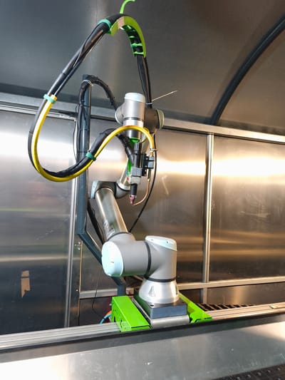 The image shows a robotic arm with a welding tool attached, positioned inside a manufacturing or industrial setting. The robotic arm is mounted on a green base and appears to be connected to various cables and hoses. The background consists of metallic walls and ceiling, suggesting a cleanroom or a specialized production area. The overall scene depicts automated industrial equipment. This specific robotic arm is located in the manufacturing facility of KUKA Roboter GmbH in Bad Honnef, Germany.
