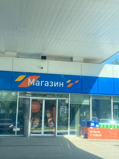 The image shows the exterior of a convenience store at a gas station. The store has a large blue sign that reads "Магазин" (Store) in white letters with an orange and yellow logo. The store entrance is made of glass doors, with a display inside showing coffee cups and a hot dog. There are also blue containers and green bottles displayed outside the store. In the reflection of the glass, a red car and trees are visible. The gas station canopy is visible overhead, made of white panels. The location is identified as Lomonosov, Russia. There are no people clearly visible in the image.