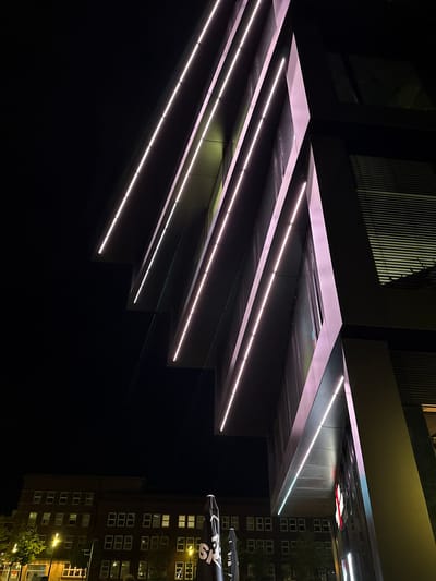 This low-angle, nighttime photograph captures the exterior of a modern building with a distinctive angular design, illuminated by white and pinkish LED strip lights. The building features repeating diagonal elements, creating a sense of depth and movement. Adjacent to this structure, a darker brick building with numerous lit windows is visible, suggesting a mixed urban environment. The scene appears to be located in the city of Duisburg, Germany, as indicated by the signage on a black umbrella in the foreground, which reads "SK"-logo and "SKLÄLLEN". The overall atmosphere is sleek and contemporary, with the lighting playing a significant role in defining the architectural features against the dark night sky. No people are clearly visible in the frame.