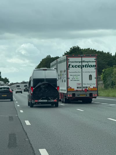 In a highway setting, under an overcast sky, a large white truck with red trim is seen from behind. The truck displays text including "Tractorpullingteam Exception" and other smaller print indicating it runs on GTL with up to 90% CO2 reduction. A white van with a black cover over what appears to be bicycles is positioned to the left of the truck. Both vehicles are traveling on a multi-lane asphalt highway, marked with white dashed lines. Other vehicles are visible further down the road. Trees and greenery line the side of the highway. The license plate on the van reads "TL-005-N" and the license plate on the truck is "36-WD-KP". The overall scene depicts road transportation.