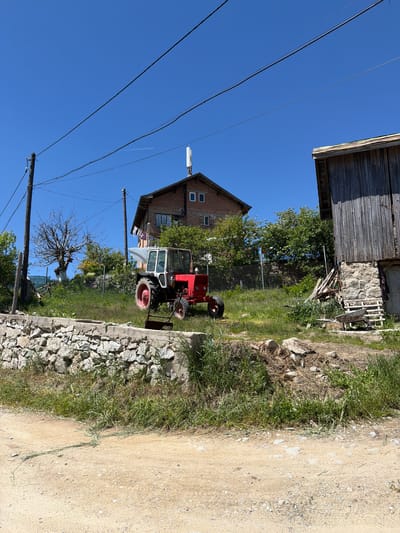 The image depicts a rural scene, possibly in Avramovo, Bulgaria. A red and white tractor is parked on a grassy area next to a stone wall and a dirt road. A brick building with windows and what appears to be a satellite dish on the roof is partially visible. A wooden structure, possibly a barn, is at the top of the image. Power lines run across the blue sky. A bare tree and other vegetation are scattered throughout the scene.