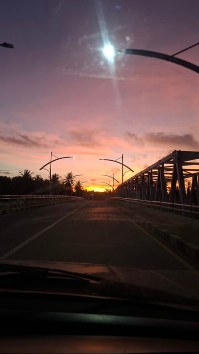 This is a photograph taken from inside a vehicle, looking forward towards a bridge at sunset. The sky is a gradient of colors, transitioning from deep purple at the top to vibrant pink and orange near the horizon where the sun is setting. Several streetlights with curved arms are visible along the sides of the road leading onto the bridge. The bridge itself is a metal truss structure, and on either side of the road, there are railings and a line of curbs. Palm trees and other foliage line the sides of the road before the bridge. The foreground shows the windshield and dashboard of the vehicle, with the wipers visible. The road is asphalt with yellow lane markings. There are no people clearly visible in the scene, though a faint indication of a motorcyclist might be present in the distance on the bridge. The overall impression is of a journey on a road in Neuboknaleung, Indonesia, at dusk.