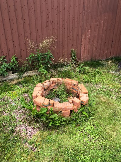 In an outdoor scene within Московский Ветеран Снт, Russia, a partially constructed ring of red bricks lies on a patch of grass and gravel. The bricks are stacked in a circular fashion, forming a boundary for what appears to be a fire pit. The surrounding area is overgrown with green grass, weeds, and some taller, dry grasses. In the background, a tall, corrugated metal fence, painted a dark reddish-brown, stands vertically. The fence has numerous small, dark, irregular marks on its surface. Several green plants are growing at the base of the fence, interspersed with the taller grasses. The lighting suggests it is daytime, with possibly overcast conditions, as indicated by the relatively diffused light. There are no people visible in the image.