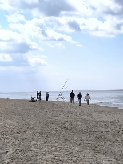 The image depicts a coastal scene, likely a beach, under a sky with scattered clouds. In the mid-ground, there's a group of approximately five individuals standing near fishing equipment, including fishing rods and a cart. To the right of this group, three people are walking along the shoreline, closer to the viewer. The environment consists of a sandy beach and the ocean. The location is likely Zandvoort aan Zee.