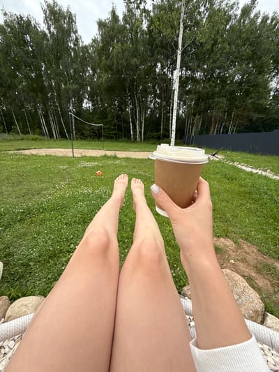 The image shows a first-person perspective of someone sitting outdoors, holding a disposable coffee cup in their right hand. Their legs are extended, and their bare feet are visible in the foreground. The background features a grassy area with scattered white flowers, a volleyball net, a sand court, and a line of birch trees behind a dark fence. The sky is overcast. The location is identified as Ryzhkovo, Russia.
