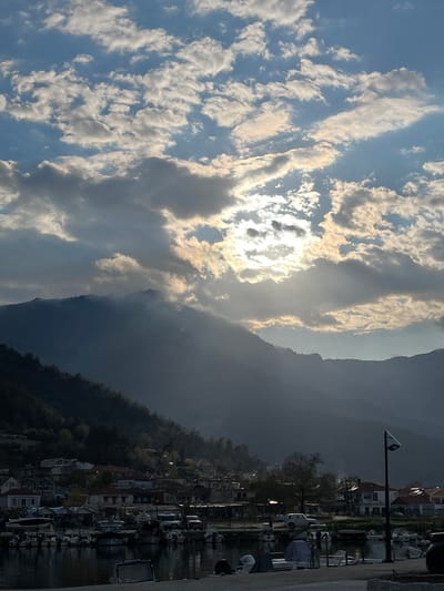 The image depicts a waterfront view in Skala Potamias, Greece. The sky is filled with white and gray clouds partially obscuring the sun. Mountain ranges are visible in the center of the frame, enveloped in a hazy atmosphere. Adjacent to the waterfront, there's a row of closely situated buildings, likely residences or businesses, leading up to a forested area. A lamp post is visible along the waterfront. Boats are moored in the water along the edge. The overall lighting suggests either early morning or late afternoon.