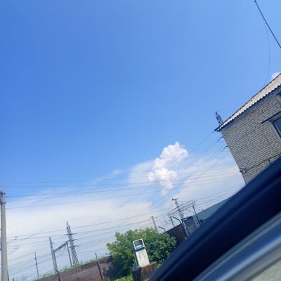 The image, captured from a vehicle in Petukhovo, Russia, shows a bright blue sky with a few wispy clouds. In the foreground, a portion of a car's interior is visible, with dark blue trim running diagonally across the bottom right of the frame. Beyond the car, a brick building with a corrugated roof is present on the right side, featuring overhead power lines. Several utility poles and electrical substations are scattered throughout the mid-ground. In the left-mid ground, a solitary, weathered green and white gas pump stands near a brick wall and dense green foliage. The overall scene suggests an outdoor, industrial or semi-urban environment under a clear day.