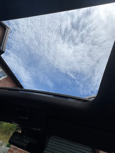 An outdoor, low-angle view captured through an open sunroof reveals a bright blue sky filled with scattered, wispy white clouds. A portion of a red brick house with a tiled roof and a window is visible on the left side of the frame. The interior of a vehicle is in the foreground, showing the headliner with controls and a rearview mirror. The overall scene suggests a clear day in Dromore, United Kingdom.