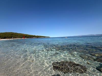 The image depicts a tranquil beach scene on a clear, sunny day. The foreground is dominated by shallow, crystal-clear turquoise water revealing a pebbled seabed. Sunlight penetrates the water, creating shimmering patterns on the bottom. In the mid-ground, a rocky shoreline meets the water, backed by a dense, green forested hill. A bright red buoy or marker stands prominently on the shore. Further out in the sea, a white rope with buoys delineates a swimming area. Several people are visible on the beach and in the water, some appearing to be sunbathing or swimming. In the distance, the coastline of what appears to be another island or the mainland is visible, featuring a mountainous landscape under a vast, cloudless blue sky. The location is identified as Općina Postira, Croatia.