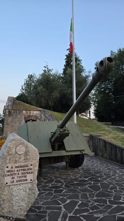 The image depicts an outdoor scene in Peschiera del Garda, Italy, featuring a historical artillery piece as the central object. The cannon, painted olive green, is positioned in a park-like setting with a stone-paved walkway and lush greenery. The Italian flag, green, white, and red, flutters on a flagpole in the background, suggesting a public memorial or historical site. A stone plaque is placed at the base of the artillery piece, bearing an inscription in Italian. The plaque reads: "A MEMORIA DEGLI ARTIGLIERI CADUTI E DISPERSI DI TUTTE LE GUERRE" which translates to "In memory of the artillerymen fallen and missing from all wars." The inscription is accompanied by a circular emblem. The scene is illuminated by natural light, indicating daytime, and the sky is clear and blue, suggesting fair weather. The overall mood conveyed is solemn and commemorative, honoring fallen soldiers. The cannon itself appears to be well-maintained, serving as a stark reminder of past conflicts. The stone wall and paving stones add to the historical ambiance of the location.