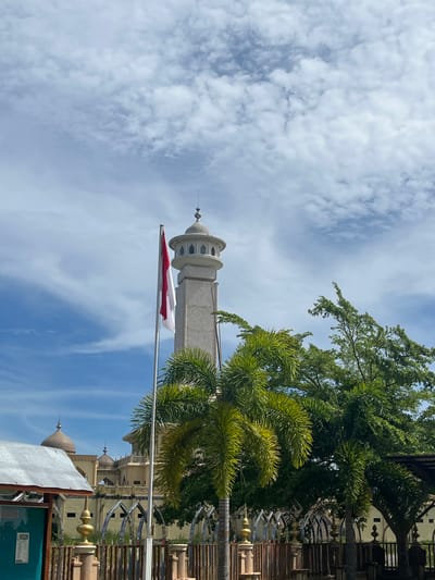 The image captures a view of a mosque minaret and surrounding architecture in Kruengmate, Indonesia, under a partially cloudy sky. A tall, light-colored minaret with a domed top and arched windows stands prominently in the center. In front of the minaret, the Indonesian flag, a bicolor of red and white, flies from a flagpole. Lush green palm trees and other foliage are visible in the foreground, partially obscuring the base of the minaret and the surrounding structures. Behind the minaret, a building with multiple domes and arched walkways can be seen. In the lower left of the frame, part of a building with a metallic roof and a green notice board is present. The scene suggests a sunny day with scattered clouds.